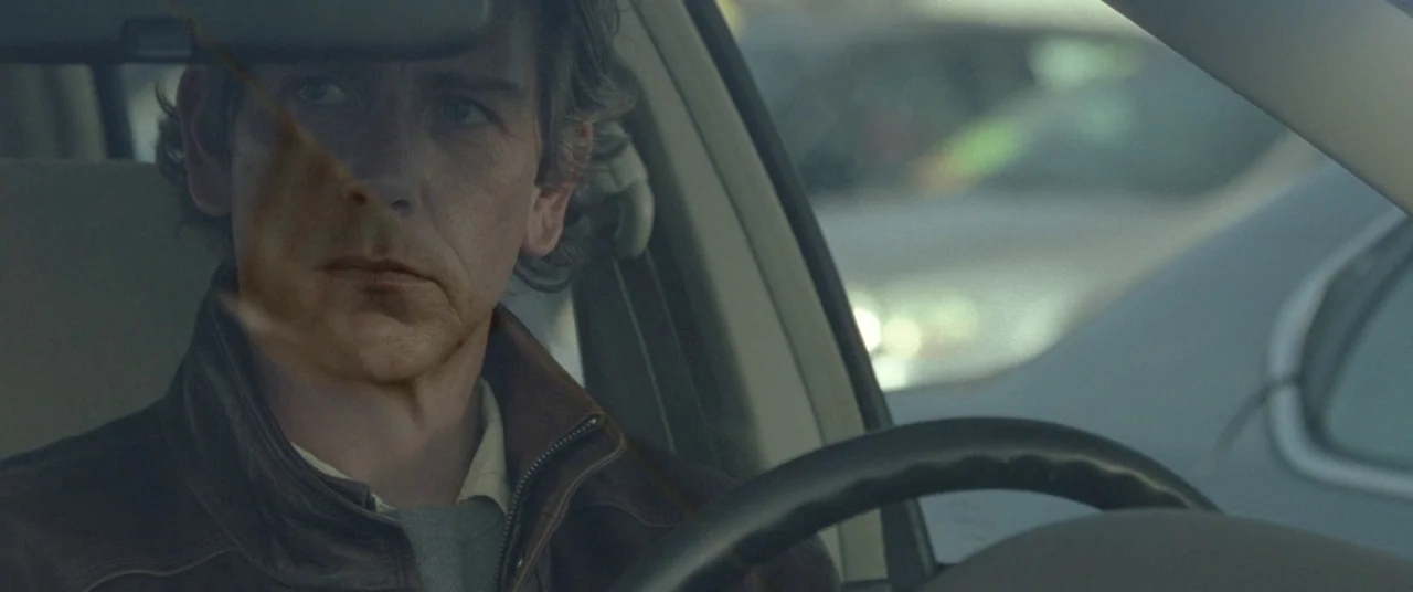Mississippi Grind (2015) movie still: closeup — The interior of a modern car shows a man seated at the wheel. The man looks ahead while resting his…