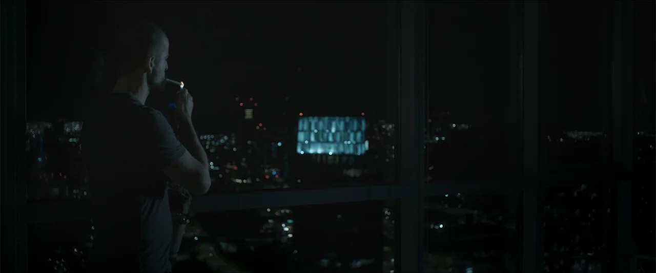 Los habitantes (2023) movie still: medium — A high-rise room with floor-to-ceiling windows overlooking a dark, illuminated city at night. A man…