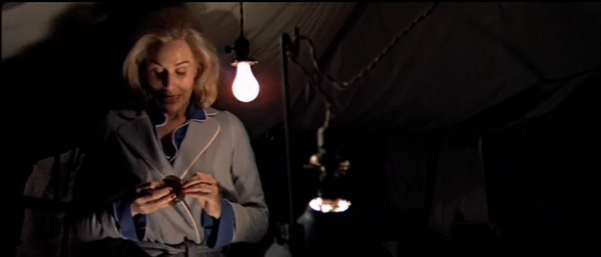 MASH (1970) movie still: medium — An interior space draped with white fabric walls and dark surroundings. A blonde woman in a grey robe examines a…