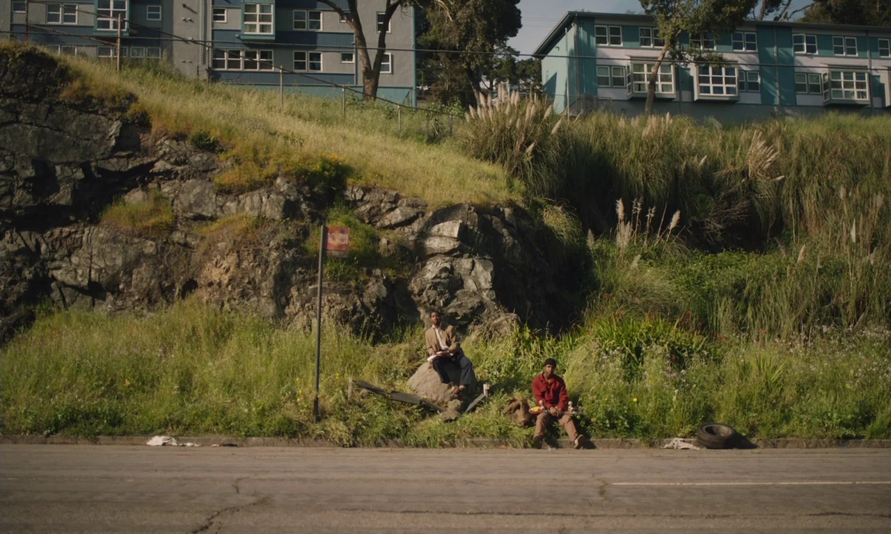 The Last Black Man in San Francisco (2019) movie still: wide — An asphalt road runs past a steep, overgrown hillside with rock formations and…