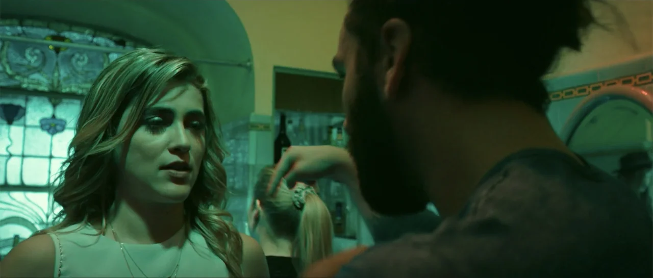 Cindy La Regia (2020) movie still: medium — A dimly lit indoor space features stained glass windows and bottles on shelves behind. A man with a dark…