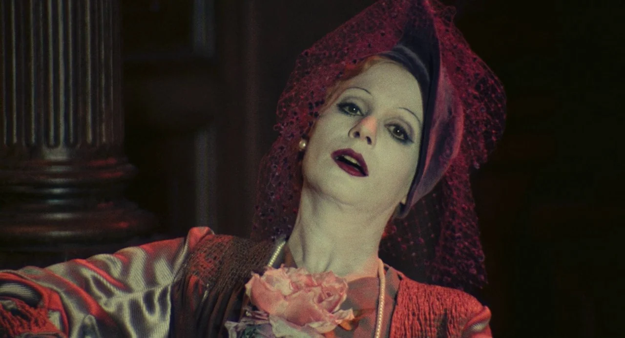 The Damned (1969) movie still: closeup — A dimly lit interior space with a dark fluted column visible to the left. An individual with pale makeup…