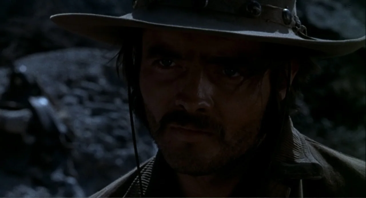 Chato's Land (1972) movie still: closeup — A rocky outdoor environment with dark, textured stone in the blurred background. A man wearing a western…