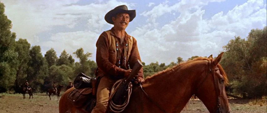 Ride Lonesome (1959) movie still: medium — A man rides a brown horse through a flat landscape filled with sparse trees. Distant figures on horses…