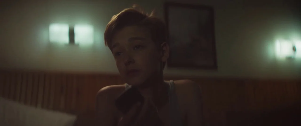 The Strange Ones (2017) movie still: medium — A dimly lit room with wall-mounted lamps and a dark picture frame behind. A young man wearing a tank…