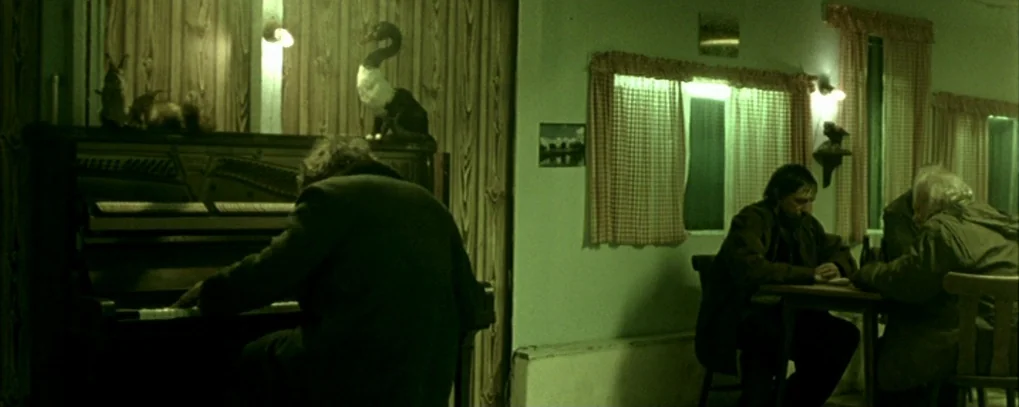 Calvaire (2004) movie still: wide — A dimly lit room features wood-paneled walls and checkered curtains over the windows. One man plays a piano…