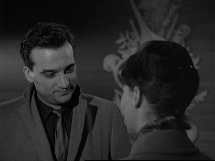 Blast Of Silence (1961) movie still: medium — A man faces a woman in a dimly lit space with a wall ornament. They appear to be in an office or…
