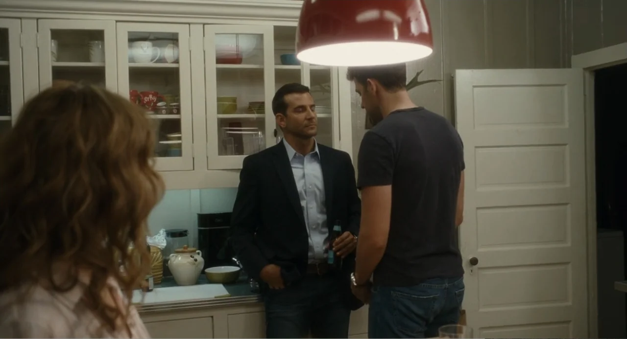 Aloha (2015) movie still: medium — A residential home kitchen with white cabinets, glass display doors, and light countertops. Two men stand in the…