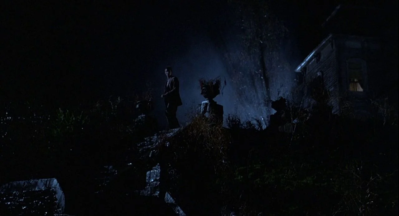 Psycho III (1986) movie still: wide — A man stands on a dark, grassy slope beneath a night sky with blue moonlight. An old two-story building with a…