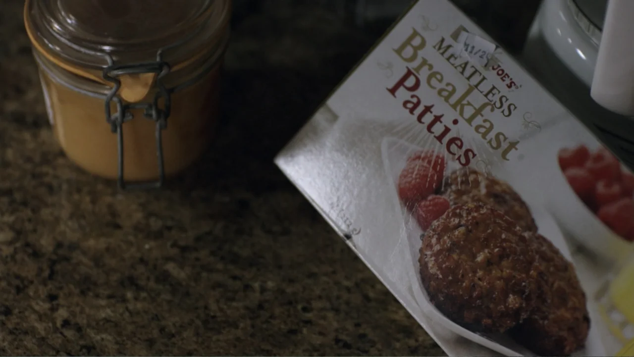 Totem (2022) movie still: closeup — A granite countertop holds a glass jar and a box of food items. A package of meatless breakfast patties rests…