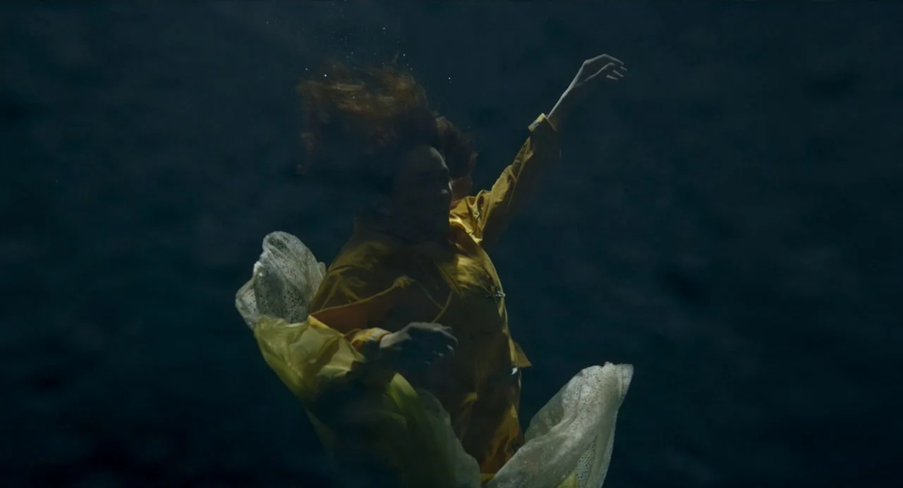 Annette (2021) movie still: medium — A person floats underwater in a vast, dark, and murky blue environment. The subject drifts with red hair fanned…