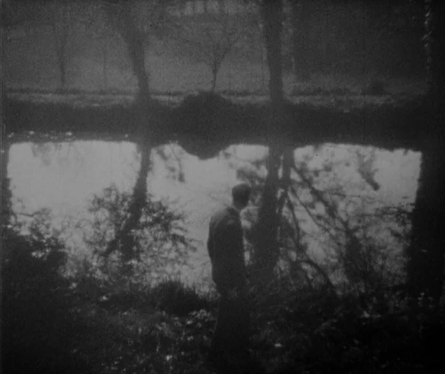 Vampyr (1932) movie still: fullbody — A dark riverbank with dense foliage frames a calm body of water reflecting trees. A man stands alone on the…