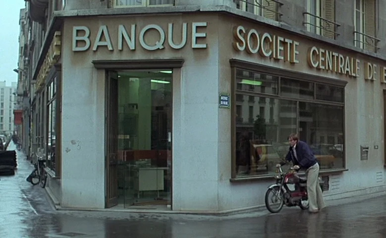 Maîtresse (1975) movie still: wide — The corner of a building features a bank entrance with large plate glass windows. A man on a red moped sits at…