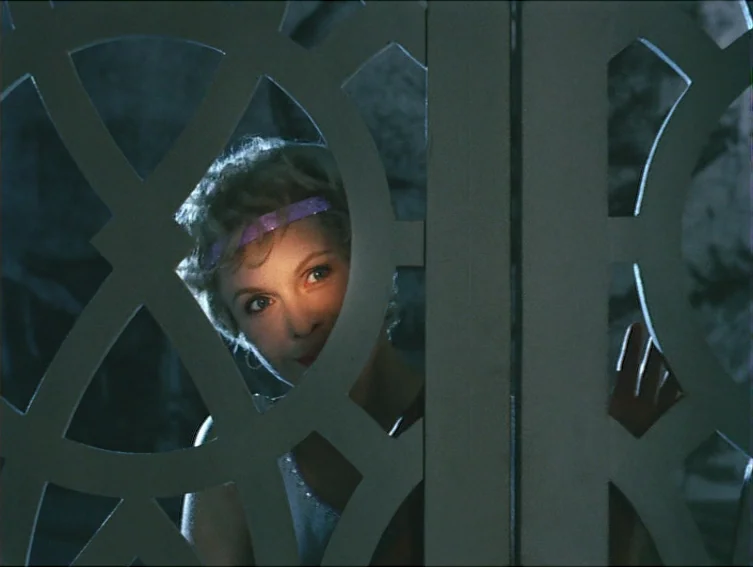 All These Women (1964) movie still: closeup — A woman looks through a cutout in a white wooden folding screen. The surrounding background is dark…