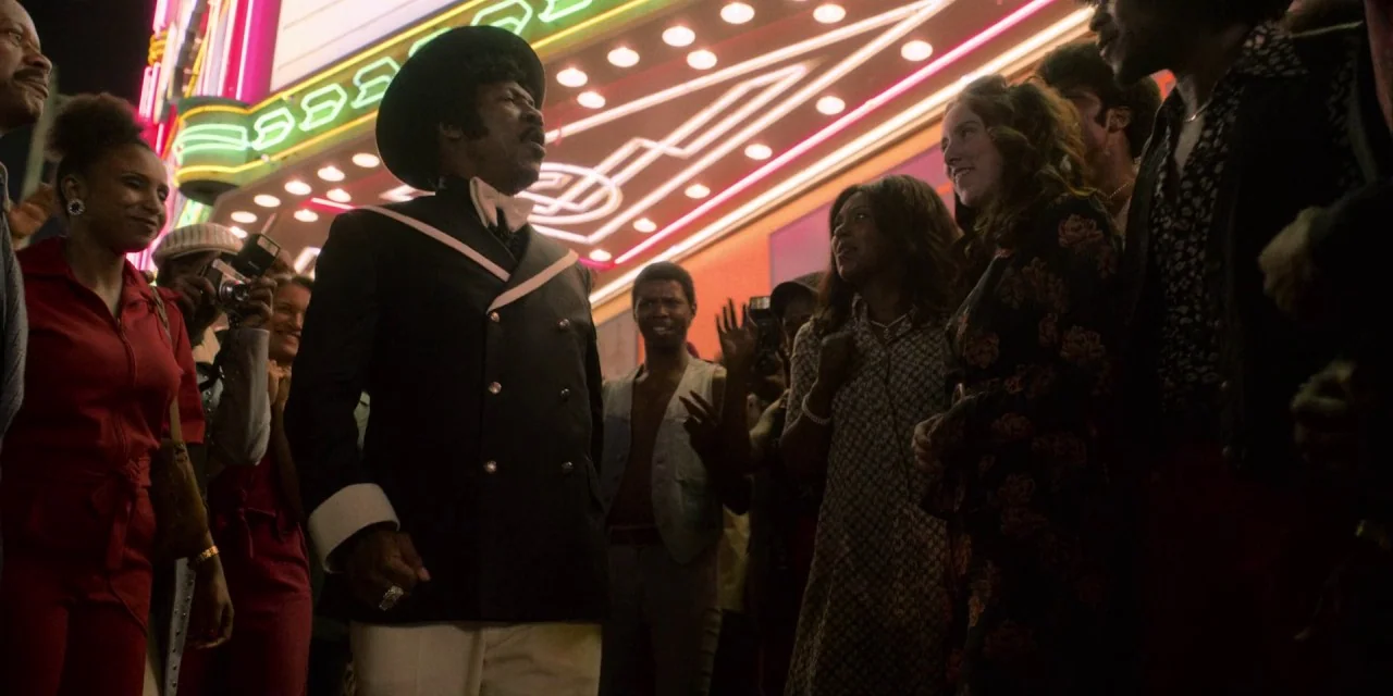 Dolemite Is My Name movie still: medium — A crowded city sidewalk at night is illuminated by the glowing marquee of a building. A man in a fedora…