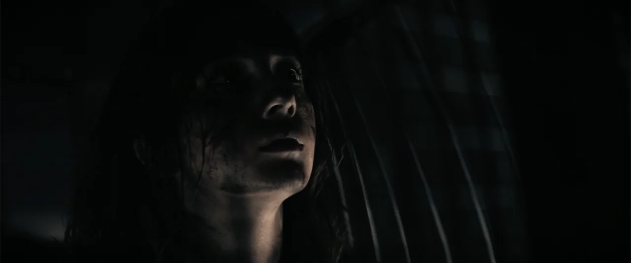 A Working Man (2025) movie still: closeup — A dimly lit interior space features dark vertical lines suggesting a grate or wall. A young woman looks…