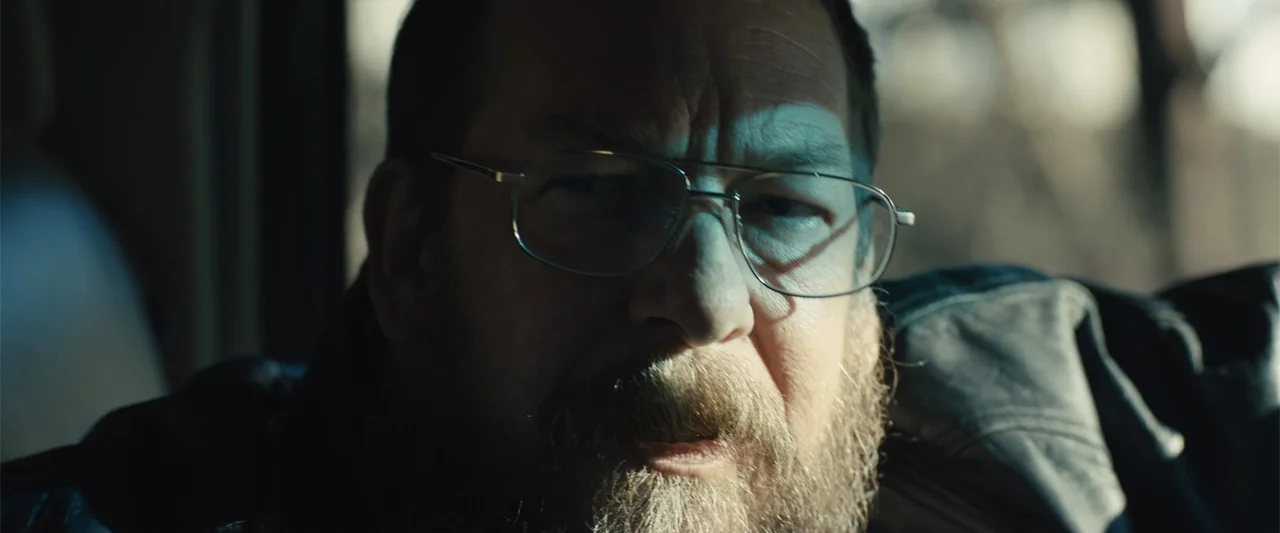 Skin (2009) movie still: closeup — A dark vehicle interior is illuminated by a single window light source. A middle-aged man with glasses and a…