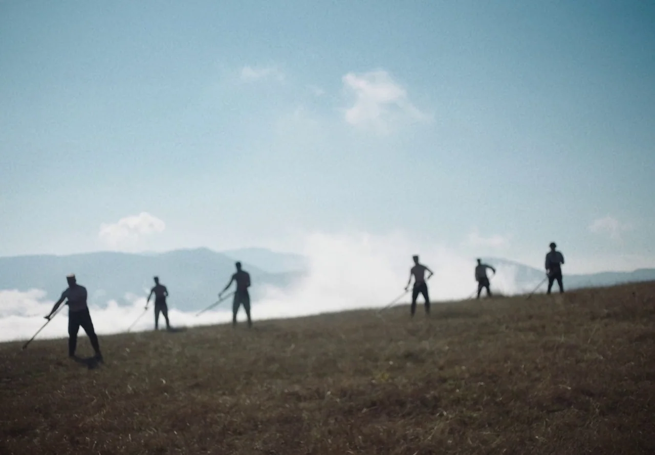 You Won’t Be Alone (2022) movie still: wide — A dry, grassy hilltop overlooks a valley filled with rolling mist under a bright sky. Six shirtless…