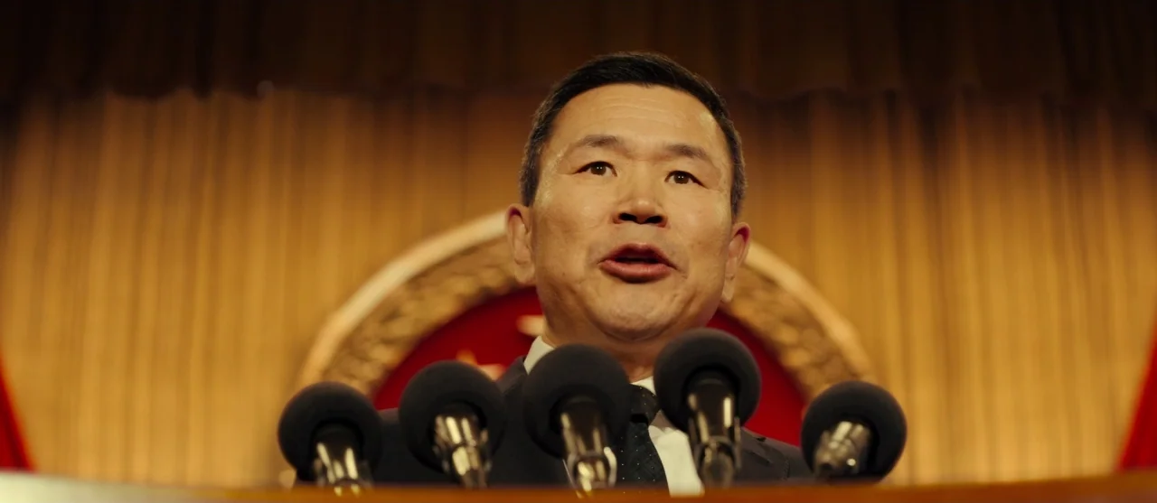 The Laundromat (2019) movie still: closeup — A man stands before a podium with a large red emblem featuring gold stars. He is positioned in front of…