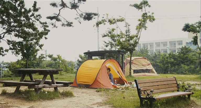 In Another Country (2012) movie still: wide — An outdoor campsite features a picnic table, a wooden bench, and two tents. A woman sits inside the…