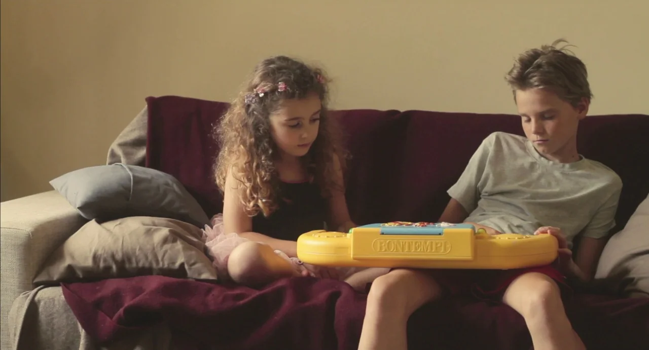 Tomboy (2011) movie still: medium — Two children sit on a maroon-covered sofa in a room with plain walls. The girl and boy play together using a…