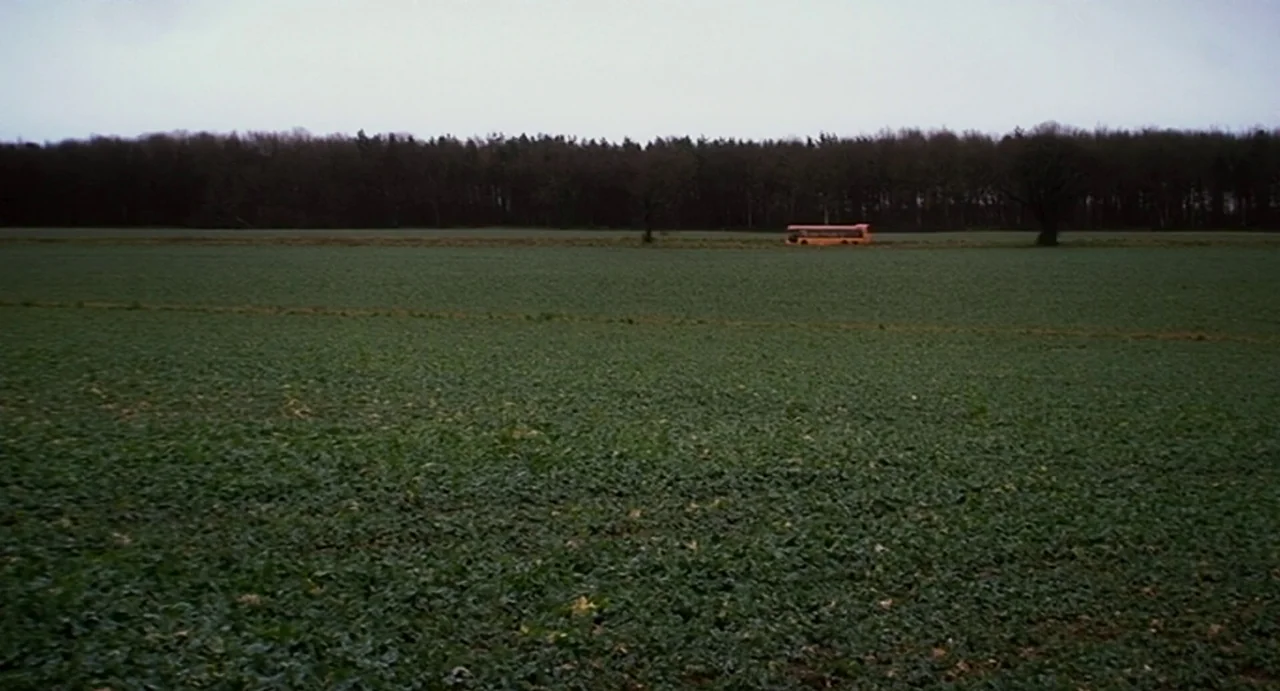Everyday (2012) movie still: establishing — A vast, flat agricultural field stretches toward a dense, dark line of trees. An orange vehicle moves…