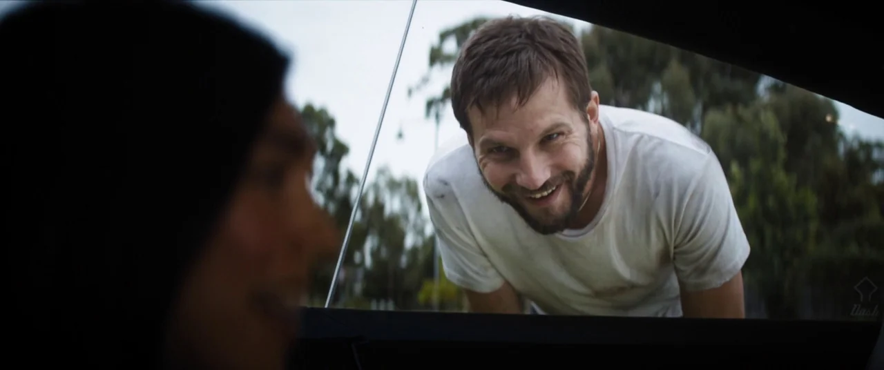 Upgrade (2018) movie still: medium — An outdoor car park with trees visible in the soft-focus blurred background. A man leans down and peers into…