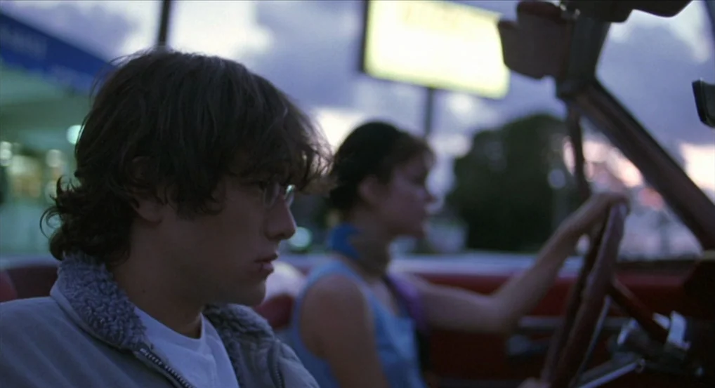 Brick (2005) movie still: medium — A man and woman sit inside a convertible car during the evening twilight hours. The woman grips the steering…