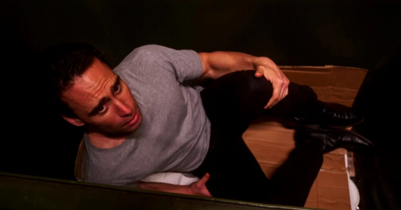T Blockers (2023) movie still: medium — A man sits inside a dark container lined with sheets of corrugated cardboard. He looks upward toward the top…