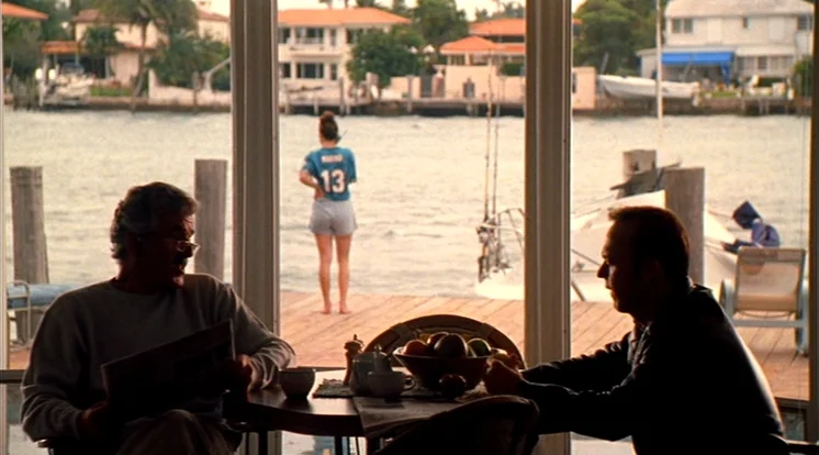 Out Of Sight (1998) movie still: medium — A man sits at a dining table facing another man across from him. A woman in a jersey stands on a wooden…