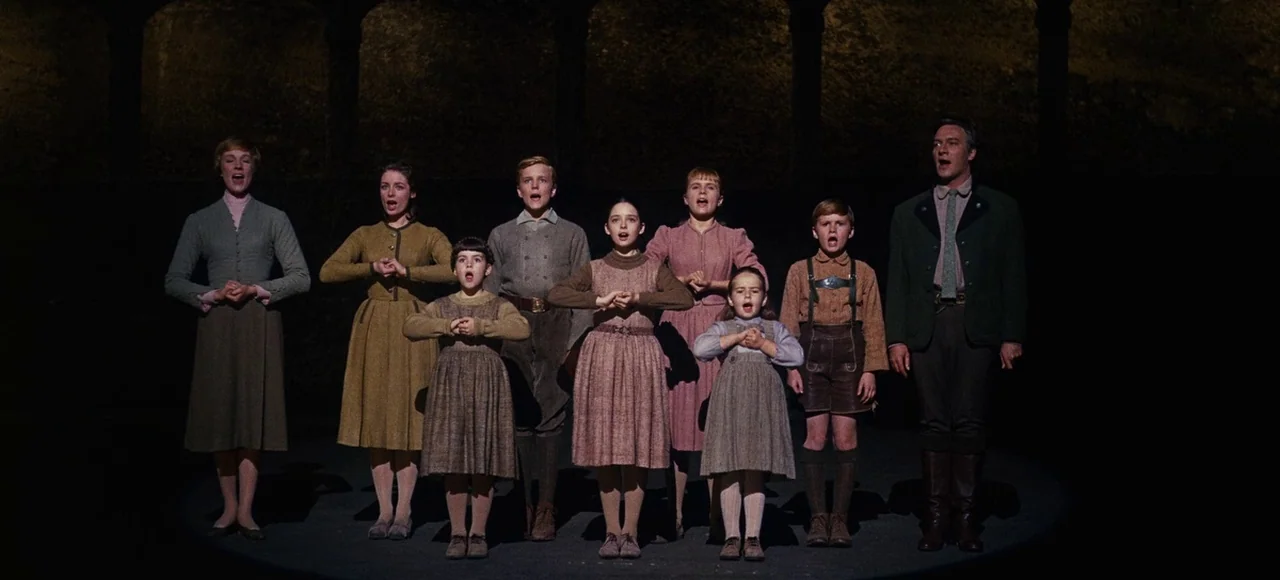 The Sound of Music (1965) movie still: fullbody — A group of eight people stand in a line on a dark outdoor stage. They sing together while…