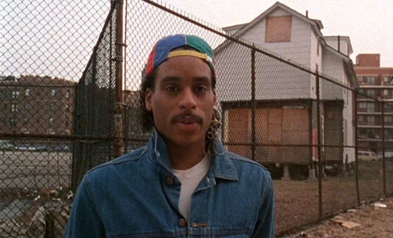 Paris Is Burning (1990) movie still: medium — An urban lot enclosed by a chain link fence features a boarded up house. A young man stands in the…