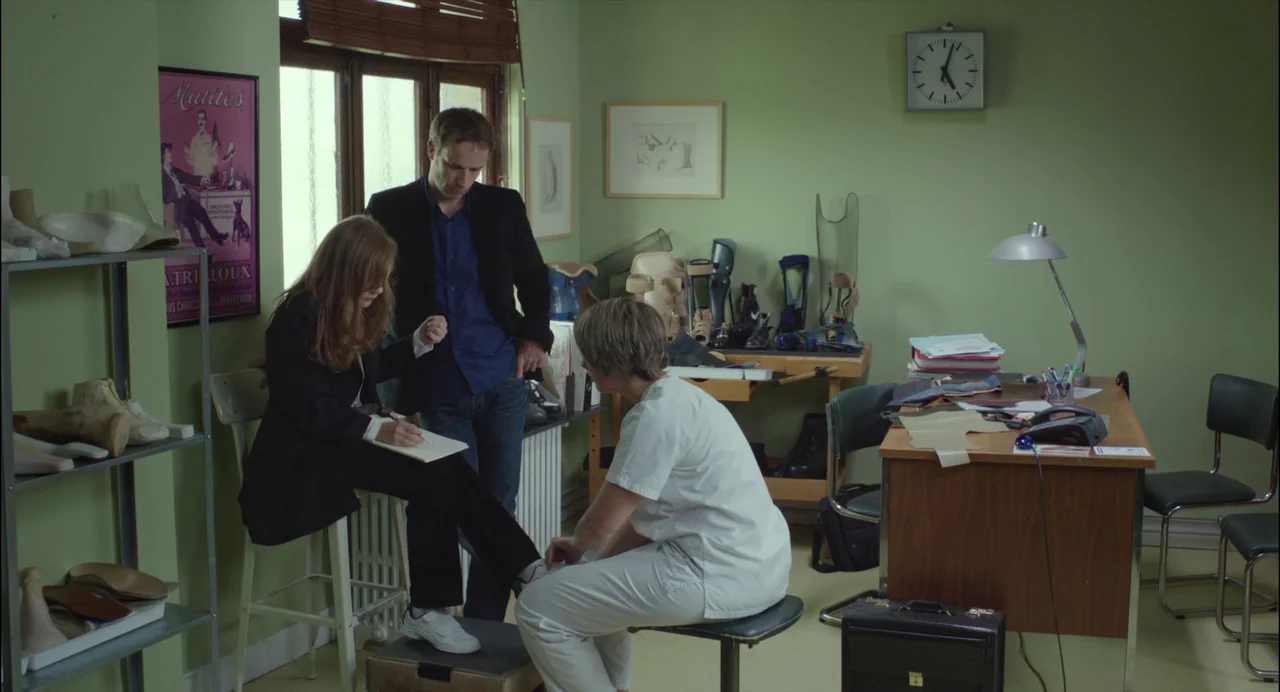 Abuse of Weakness (2013) movie still: medium — An orthopedic workspace features sage green walls, a cluttered wooden desk, and various leg braces. A…