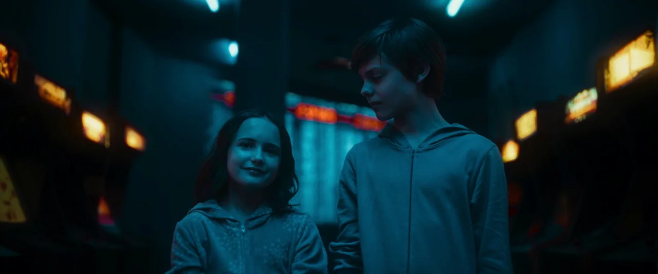 Boy Kills World (2023) movie still: medium — An dimly lit arcade interior is flanked by rows of glowing game cabinets. Two young people stand in the…
