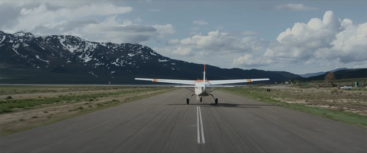 Flight Risk (2025) movie still: wide — A small light aircraft moves along an asphalt runway set within a flat valley. Snow-capped mountains stand…