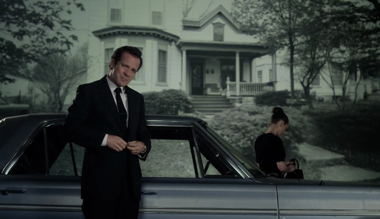 Experimenter (2015) movie still: medium — A suburban residential street with a white house in the background. A man stands in the foreground beside…
