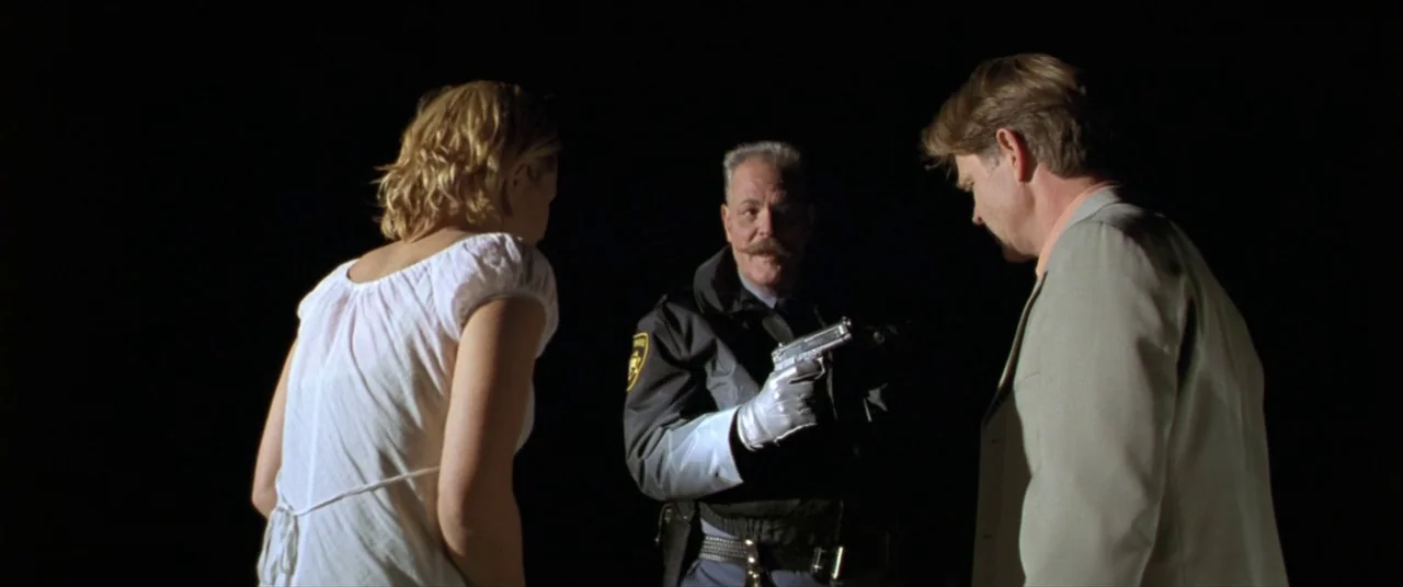 The Cooler (2003) movie still: medium — A dark night setting with no visible background environment. A uniformed police officer holds a pistol while…