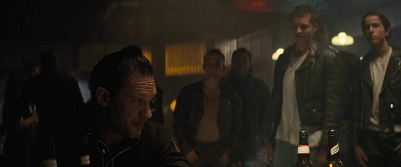 The Bikeriders (2023) movie still: medium — A dimly lit 1960s-style pub features several men gathered in a hazy, smoke-filled space. A man in a dark…