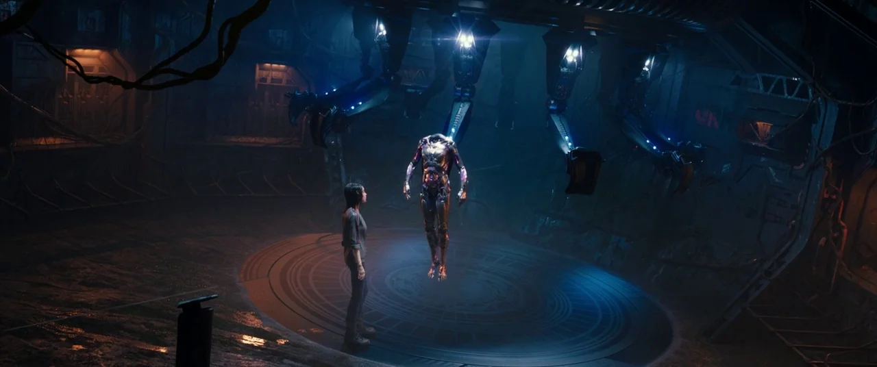 Alita: Battle Angel movie still: wide — A dark industrial chamber features a metallic floor with a glowing circular platform. A human girl observes…