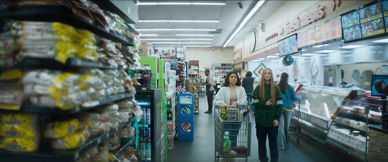 Kajillionaire (2020) movie still: medium — A well-lit grocery store aisle features metal shelves packed with diverse retail products. Two women…