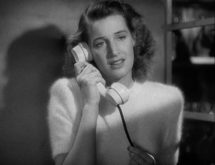 Music in Darkness (1948) movie still: closeup — A woman stands in an indoor space with a plain wall behind her. She is holding a corded telephone…