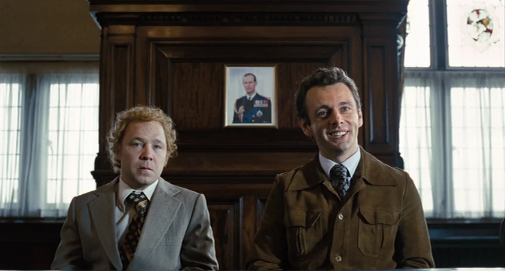 The Damned United (2009) movie still: medium — Two men sit before a dark wood paneled wall containing a small framed portrait. The subjects sit side…