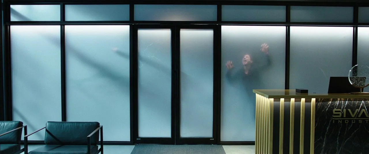 Shazam! (2019) movie still: wide — A modern corporate lobby features a reception desk and large frosted glass panels. Two silhouettes press their…