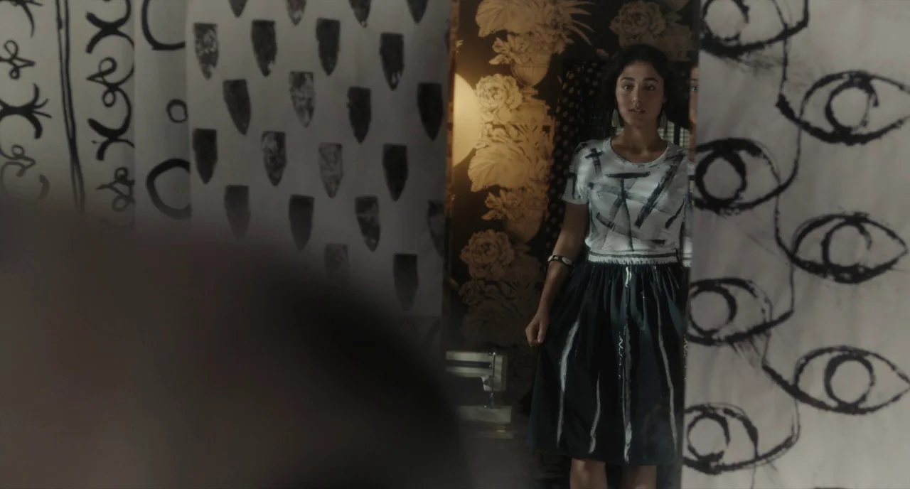 Paterson (2016) movie still: medium — A workspace features walls covered in paper printed with hand-drawn eyes and geometric shapes. A woman stands…