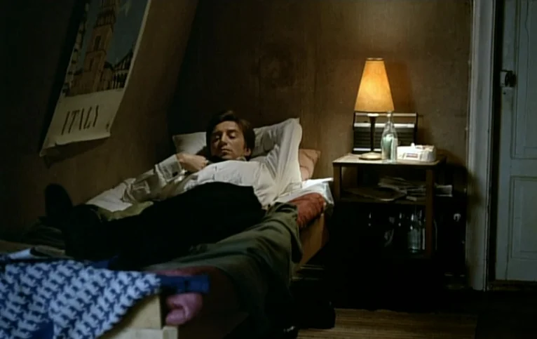 Giliap (1975) movie still: medium — A small room with wood-paneled walls and a travel poster on the sloped ceiling. A man rests on a bed near a…