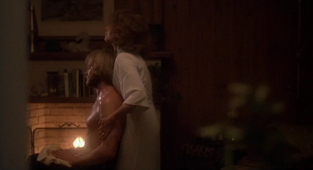 Coming Home (1978) movie still: medium — A dimly lit room features a brick fireplace with a mantle holding various books. A woman in a white…