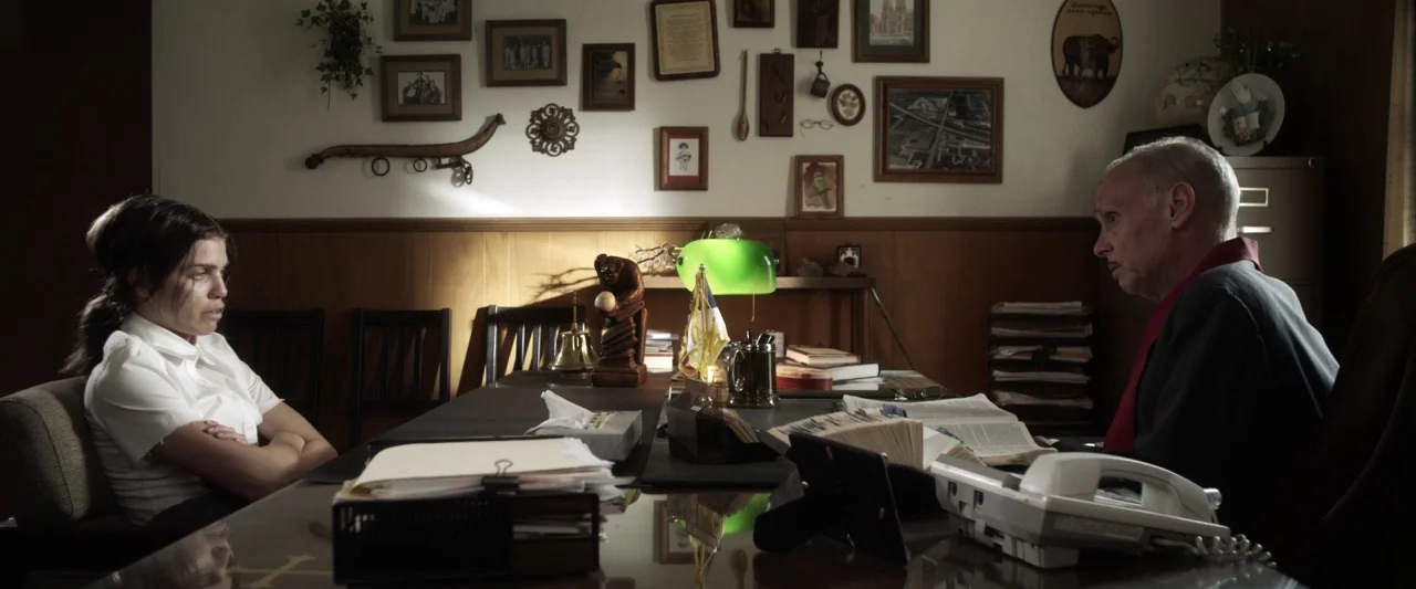 Excision (2012) movie still: medium — An office features wood-paneled walls decorated with numerous small framed pictures and wall decor. A young…