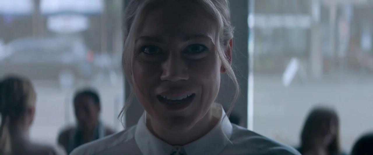 Pig (2021) movie still: closeup — An interior space with a window in the background providing ambient light. A woman wearing a white shirt stares…