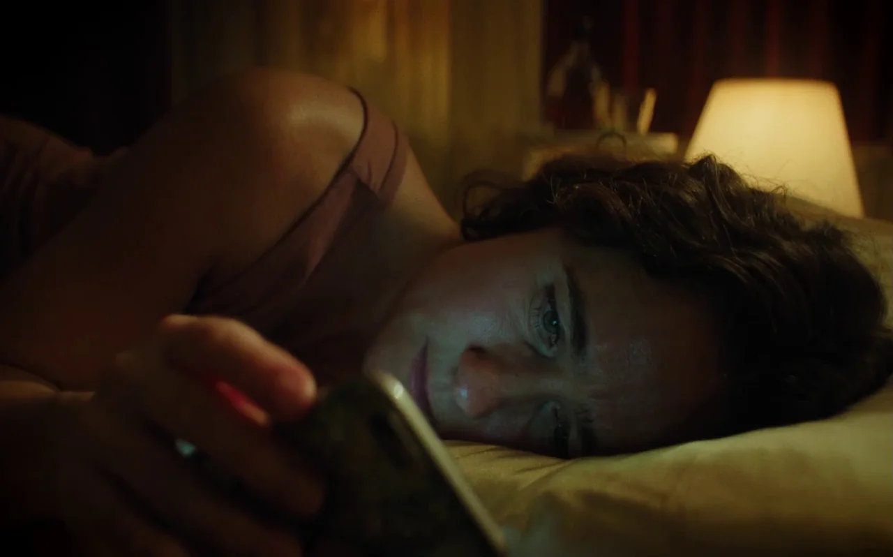 Knocking (2021) movie still: closeup — A person lies on pillows in a dark bedroom lit by a nearby bedside lamp. The subject watches a smartphone…