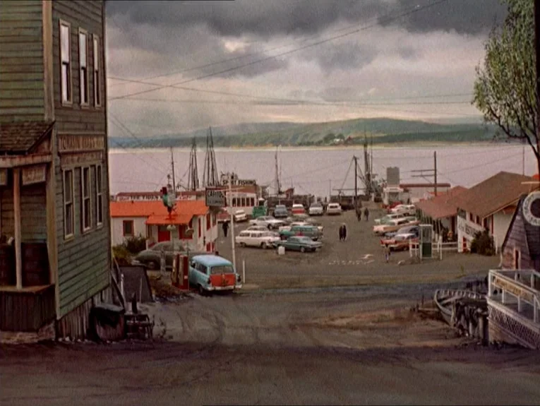 The Birds movie still: wide — A quiet coastal town waterfront features weathered wooden buildings and a parking lot. Multiple vintage cars are…
